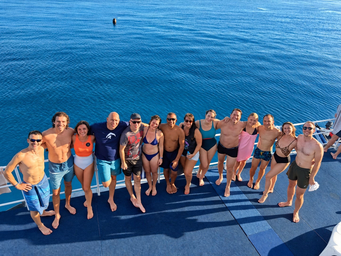 A group of divers from the Nomad Cruise community standing arm in arm on a boat deck, the open sea behind them